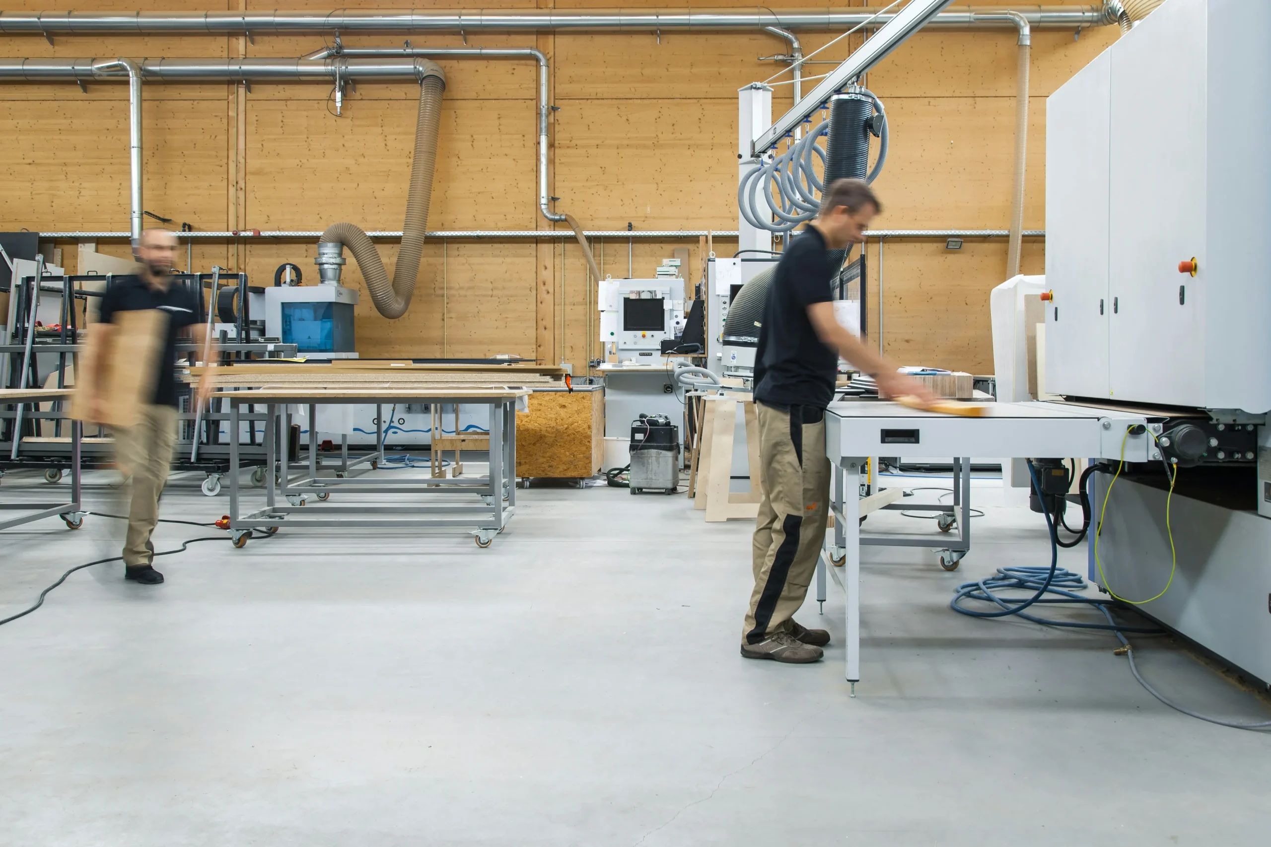 Dois homens trabalhando em uma marcenaria moderna equipada com máquinas industriais e mesas de trabalho, um deles operando uma máquina e o outro caminhando com uma tábua de madeira.